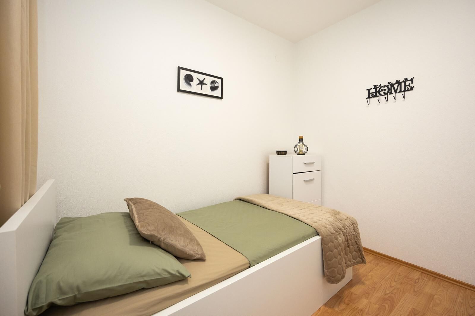 Une petite chambre avec un lit simple, une couverture verte, deux oreillers, une petite table de chevet avec un vase et une armoire blanche.