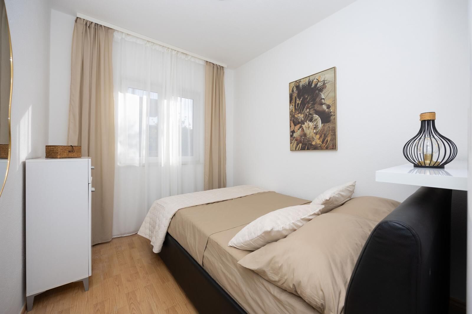 A bedroom with a bed, a dresser, a window with sheer curtains, a wall-mounted artwork, and a small table with a decorative lamp.