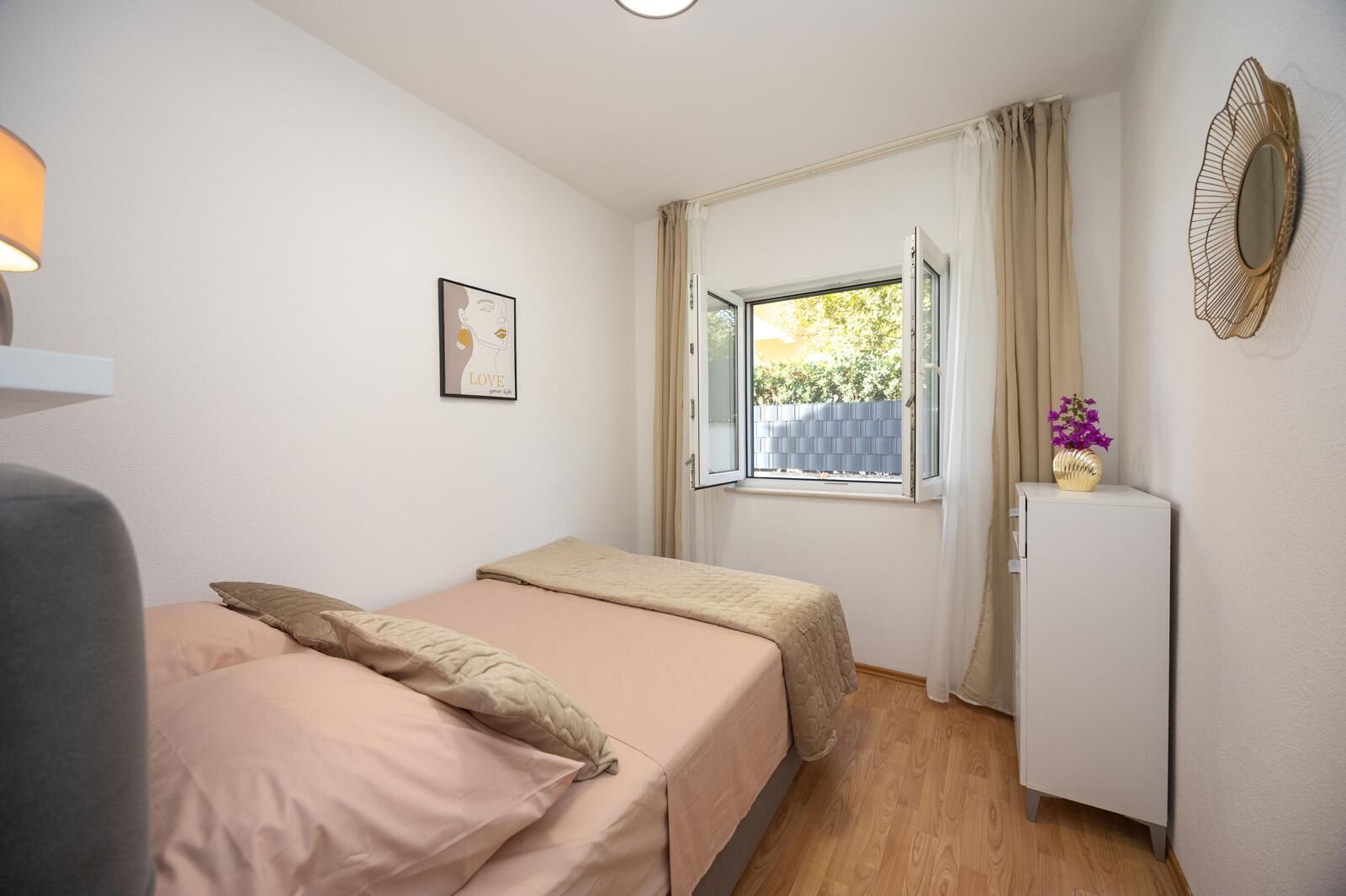 Une petite chambre à coucher, bien rangée, avec un lit, une table de chevet ornée d’un vase, une fenêtre avec des rideaux et un miroir fixé au mur.
