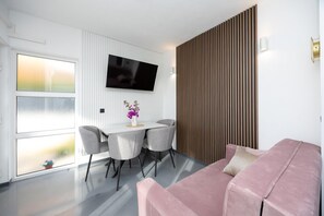Ein modernes Wohnzimmer mit einem rosafarbenen Sofa, einem Esstisch mit vier Stühlen, einem flachbildfernseher an der Wand und einem großen Fenster mit Aussicht.
