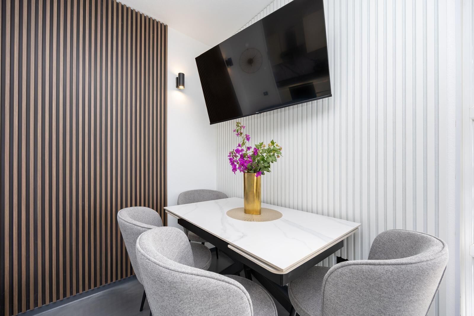 A modern conference room with a white table, four grey chairs, a gold vase with purple flowers, a mounted flat-screen TV, and a wooden slat wall.