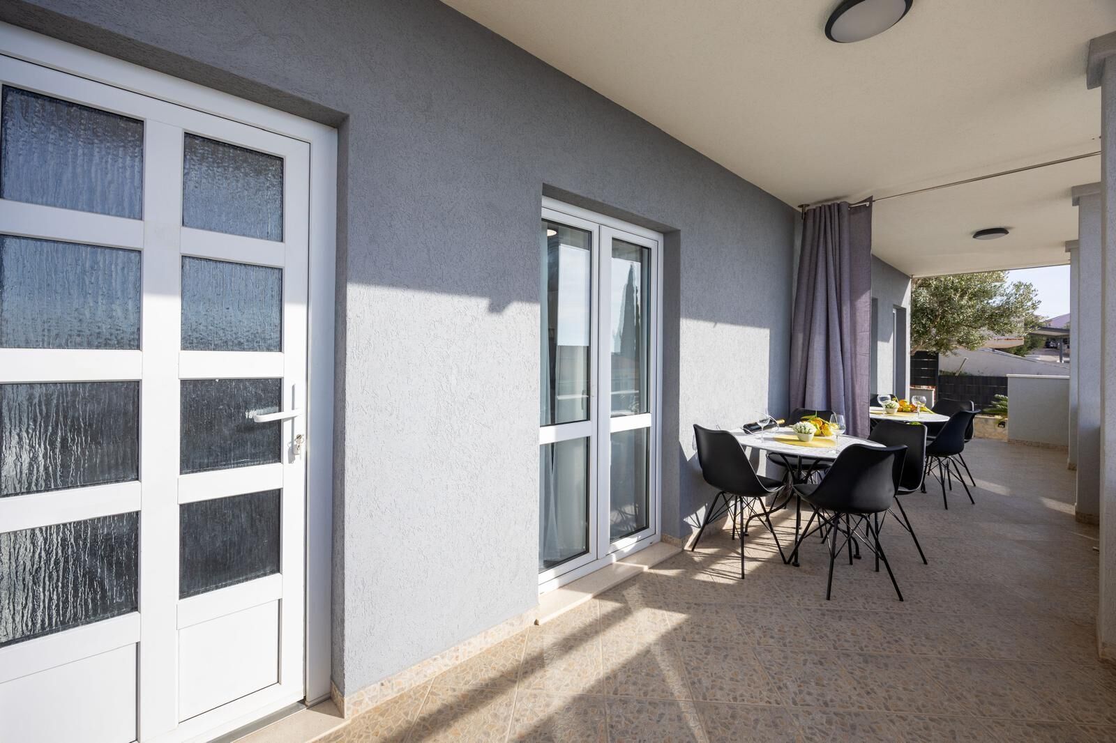 A modern outdoor dining area with a table and chairs, large windows, and a grey wall.