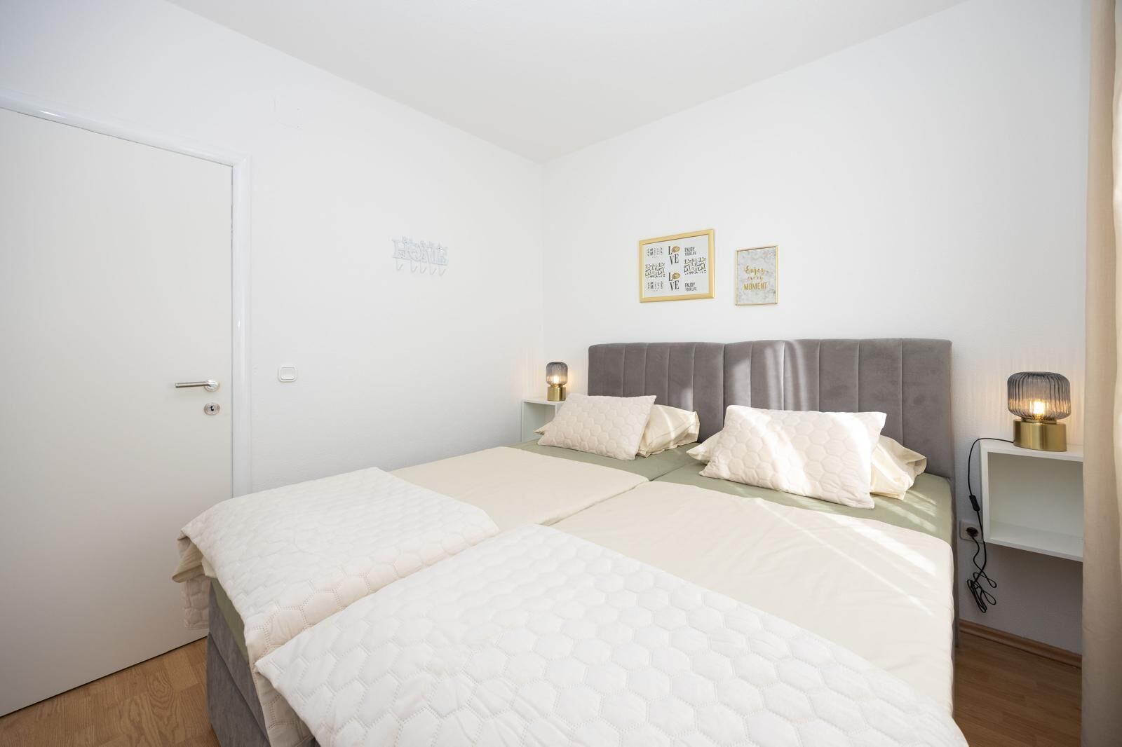 A double bed with a grey headboard, two bedside tables with lamps, and a white door.