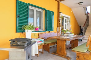 Un comedor con mesa y bancos de madera, una ventana con contraventanas verdes y una escalera.