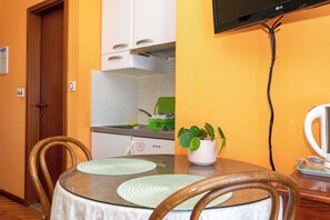 Un comedor con una mesa redonda, sillas, un televisor montado en la pared y una cocina al fondo.