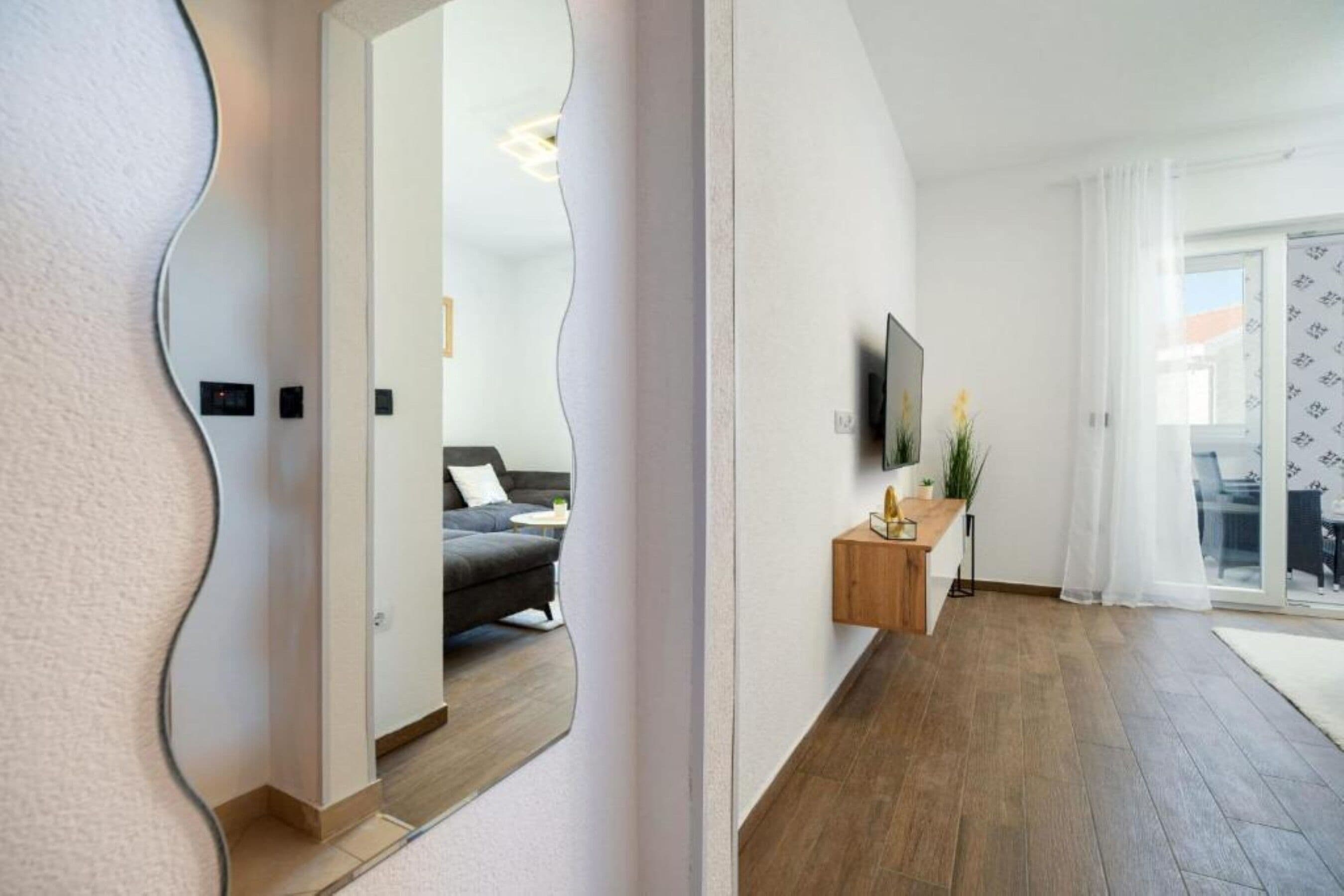 A modern interior with a wooden floor, a grey sofa, a TV on a wooden stand, and a glass door leading outside.