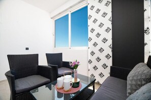 Ein Zimmer mit zwei Korbstühlen, einem Sofa, einem Glastisch mit Vase und Tassen sowie einem Fenster mit gemusterten Vorhängen.