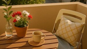 Une table ronde en bois, sur laquelle se trouvent un pot de fleurs, une tasse et une chaise agrémentée d’un coussin à motifs, le tout placé sur un balcon.