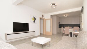 A modern living room with a flat-screen TV, a white coffee table, and a sofa. There is a dining area with a table and chairs visible in the background. - Apartments Jozak (Trogir)