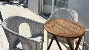 Eine Terrasse mit Korbstühlen, einem kleinen Holztisch, einer Schiebetür und einem Gebäude mit heller Fassade.