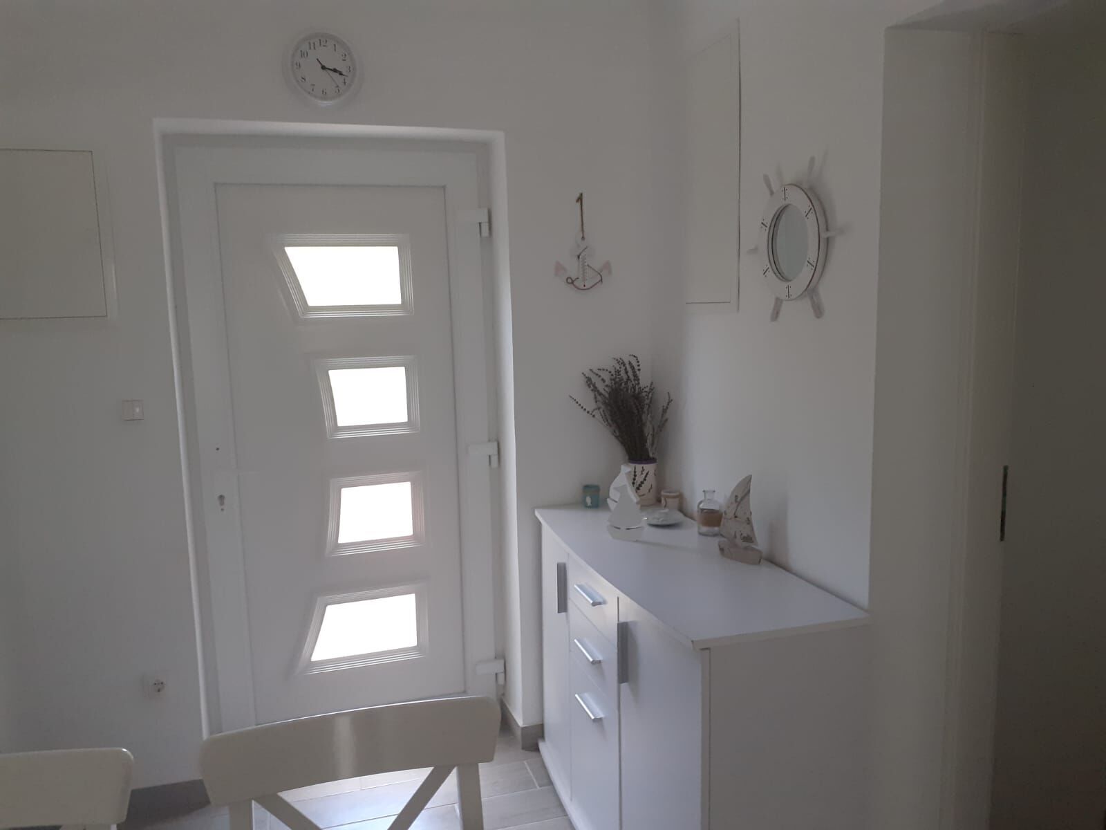 Ein Zimmer mit weißer Tür, einem kleinen Tisch mit Stuhl und einem weißen Schrank mit Pflanze darauf.