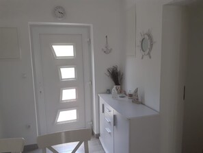 Ein Zimmer mit weißer Tür, einem kleinen Tisch mit Stuhl und einem weißen Schrank mit Pflanze darauf.