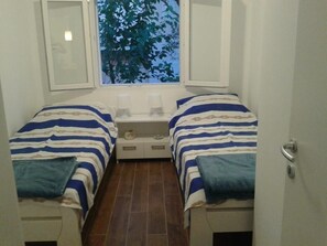 Ein kleines, kompaktes Zimmer mit zwei Einzelbetten, einem kleinen Tisch und einem Fenster mit Blick auf Bäume.