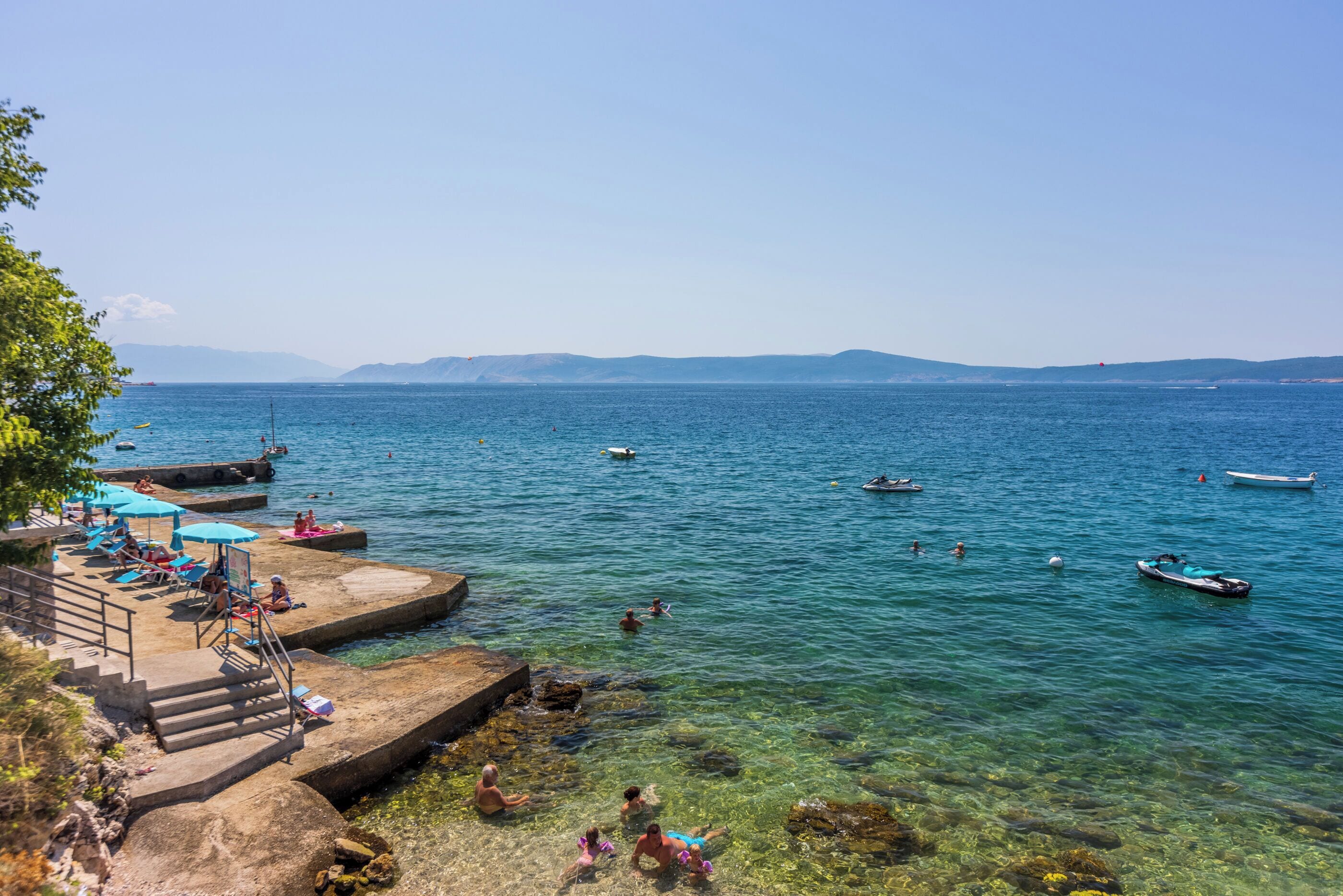 Una zona costiera con acque cristalline, scale che conducono alla spiaggia e diverse imbarcazioni sull'acqua.