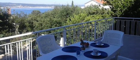 A balcony with a table set for two, overlooking a coastal landscape with trees and a distant view of the sea.