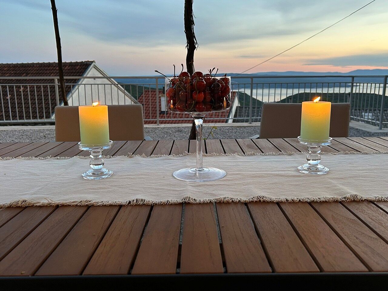 A wooden deck with two candles on a tablecloth, a glass of drink, and a bowl of cherries.