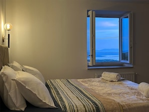 A bedroom with a bed, pillows, a bedside table, a lamp, and a window with a view of the sea. - House Martin (Vinodol)