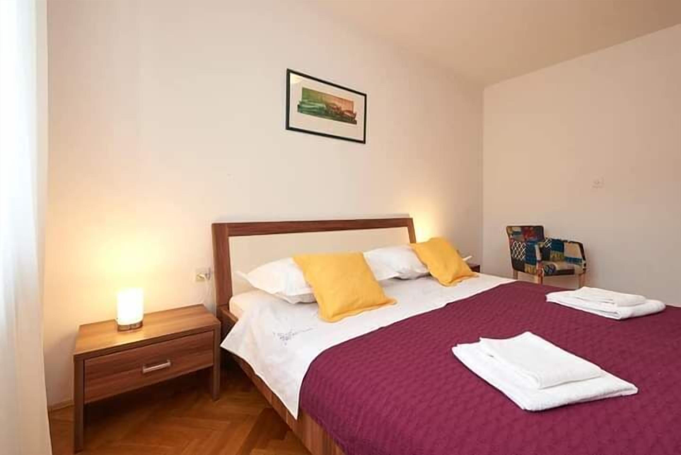 A neatly made bed with a wooden headboard, a bedside table with a lamp, and a chair with a colorful cushion.