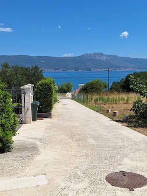Ein Kiesweg führt zu einem Tor, mit Blick auf das Meer und die Berge im Hintergrund.