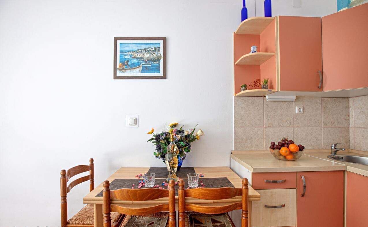 A dining table with chairs, a vase with flowers, a framed picture on the wall, and a kitchen area with cabinets and a fruit bowl.