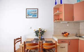 Ein Esstisch mit Stühlen, eine Vase mit Blumen, ein gerahmtes Bild an der Wand und eine Küchenzeile mit Schränken und einer Obstschale.