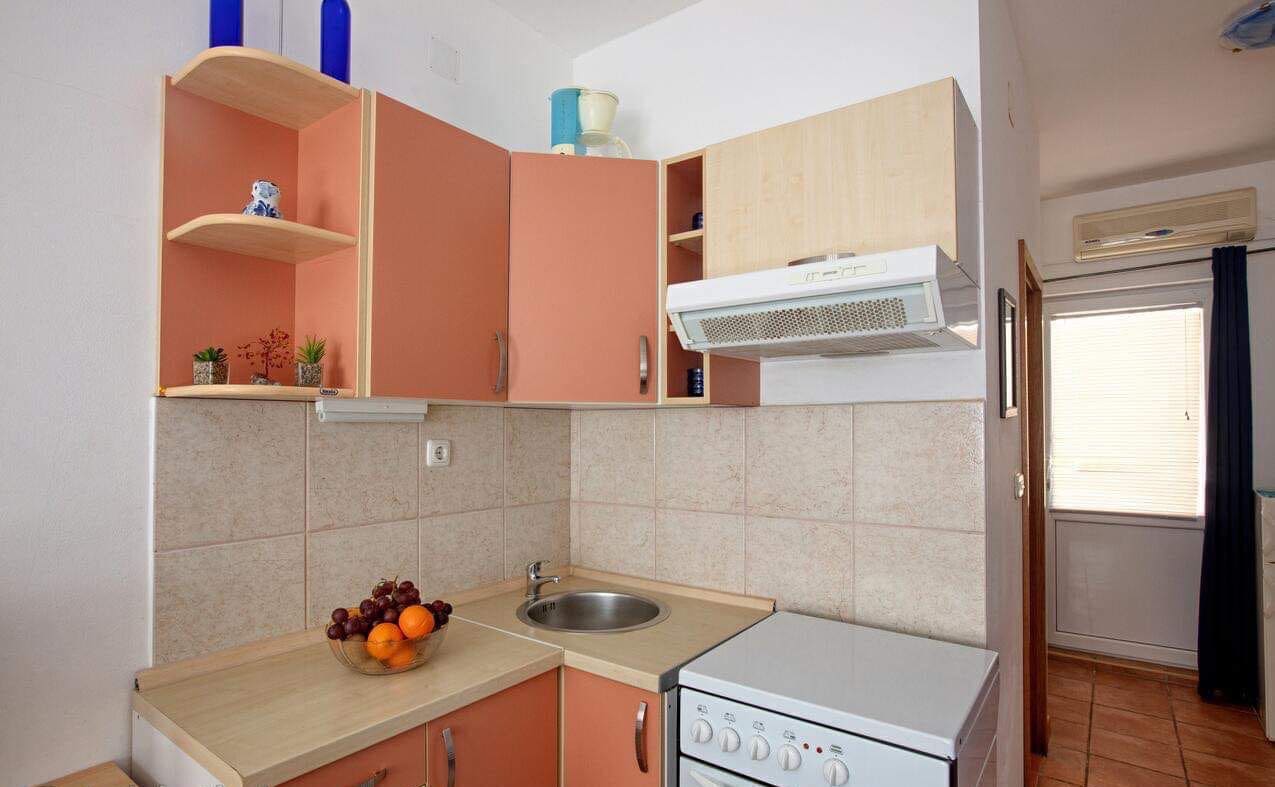 Eine kleine Küche mit Spüle, Herd und Mikrowelle. Es gibt Schränke und ein Fenster mit Jalousien.