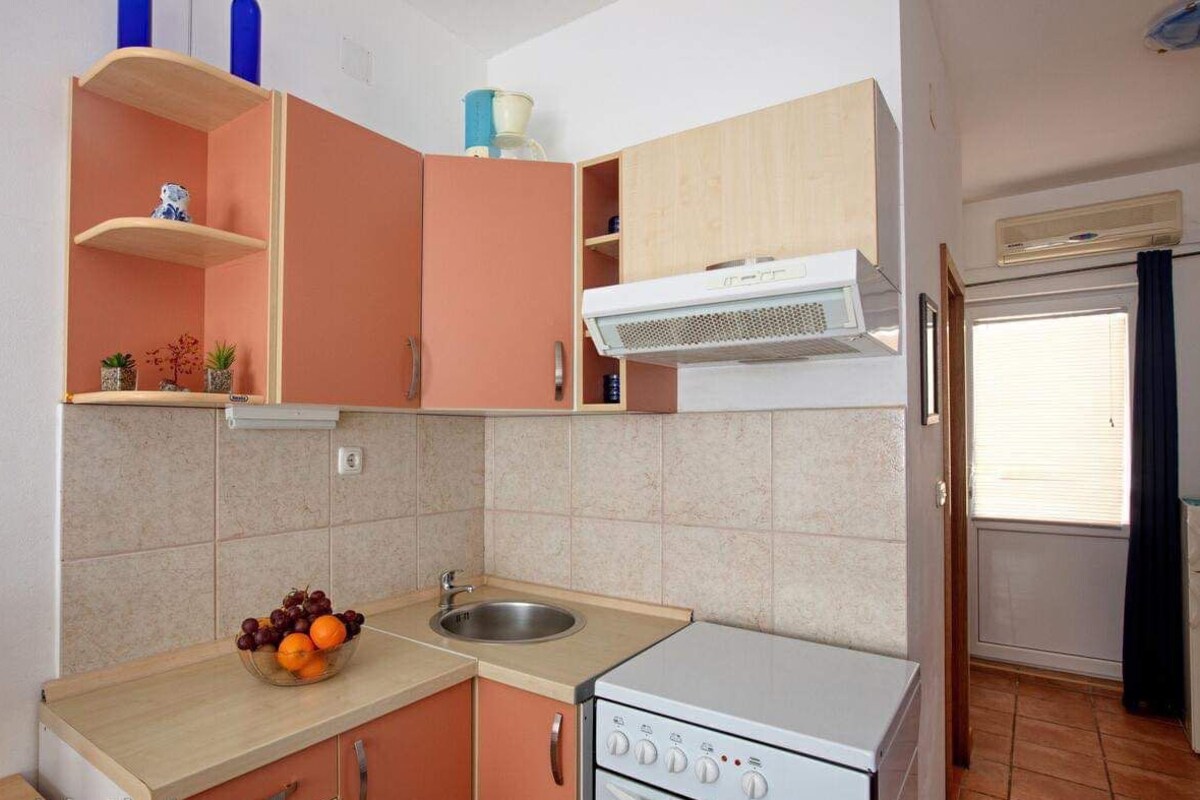 A compact kitchen with a sink, a stove, and a microwave. There are cabinets and a window with blinds.