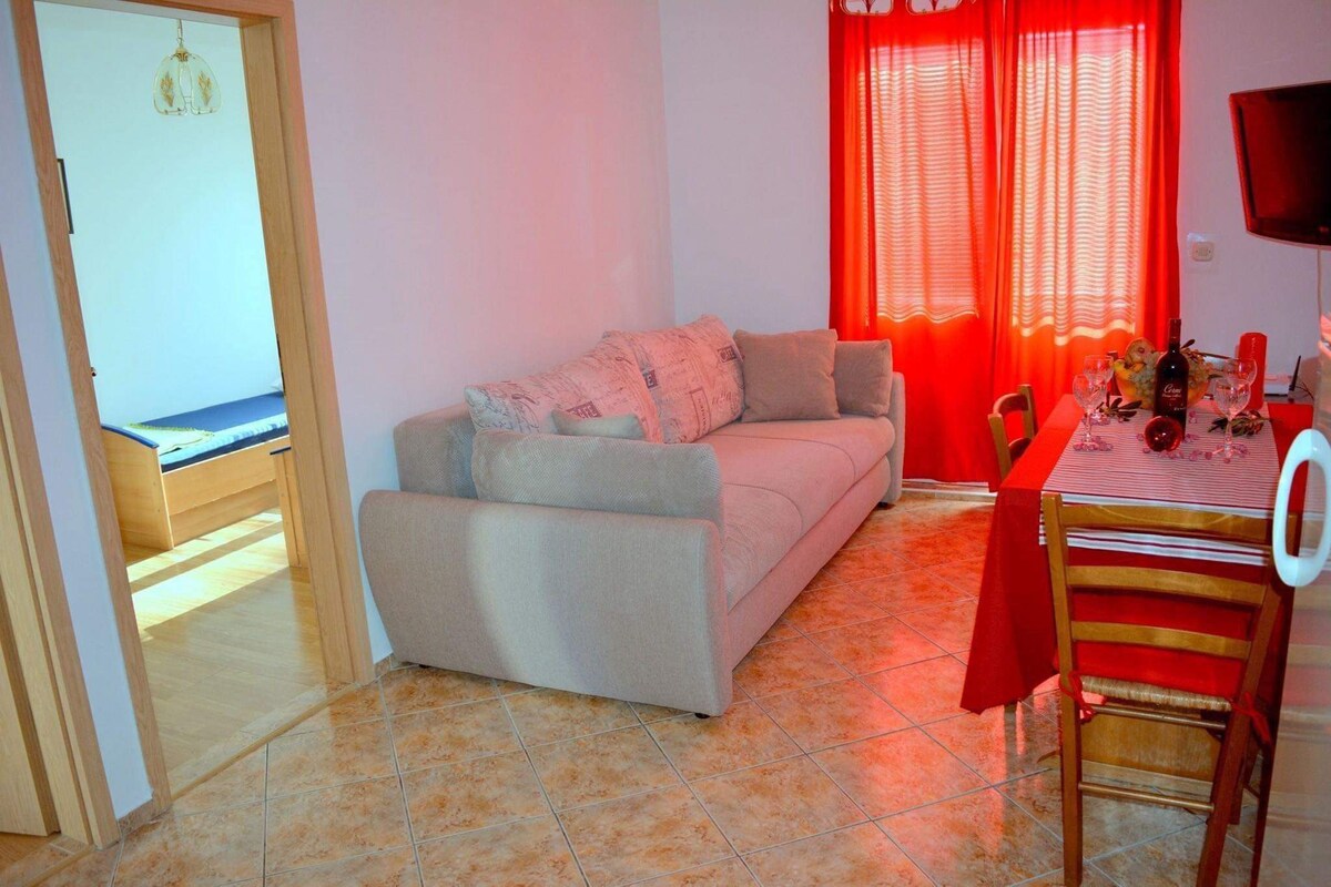 A living room with a sofa, a dining table with chairs, a TV, and a window with red curtains.