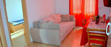 A living room with a sofa, a dining table with chairs, a TV, and a window with red curtains.