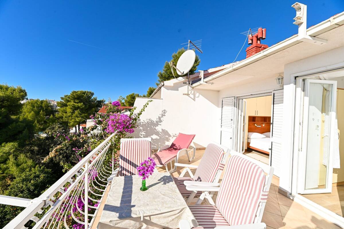 A balcony with white furniture, a table with a vase, and a view of the surrounding greenery and buildings.