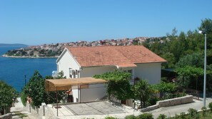 Ein Küstenhaus mit Ziegeldach, einer kleinen Veranda und Blick auf das Meer sowie eine Stadt im Hintergrund.
