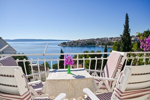 Ein Balkon mit weißen Stühlen und einem Tisch, der auf eine Küstenstadt und das Meer blickt.