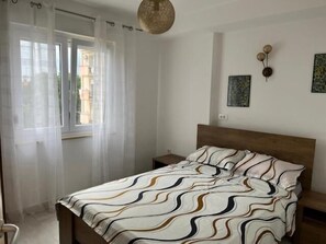 Ein Schlafzimmer mit einem Bett, einem Nachttisch, einem Fenster mit durchsichtigen Vorhängen und einer Wandleuchte.