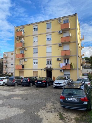 Ein mehrgeschossiges Wohngebäude mit Balkonen, Klimaanlagen und einem Parkplatz mit mehreren Autos.