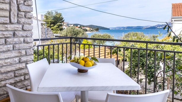 A balcony with a table and chairs, a bowl of fruit, and a view of the sea and distant buildings. - House Holiday House (Okrug)