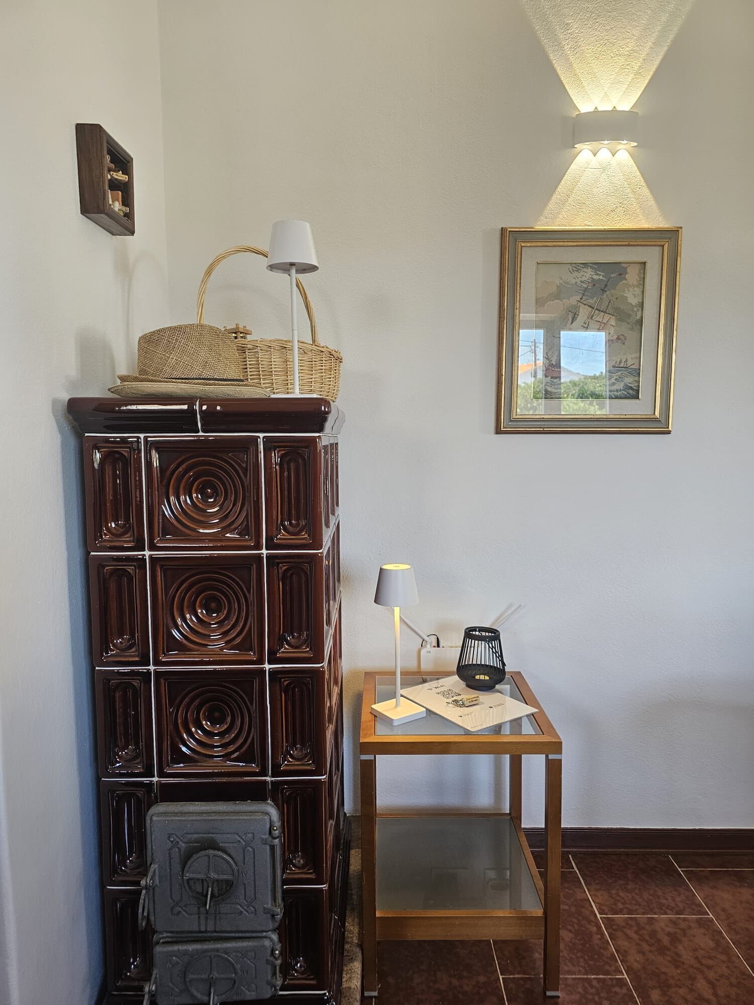 Habitació d'angle amb un armari de fusta, una taula petita amb una làmpada i un quadre encimbellat a la paret.