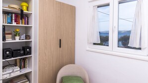 Una habitación con estantería, armario de madera, ventana con vistas y sillón con cojín.
