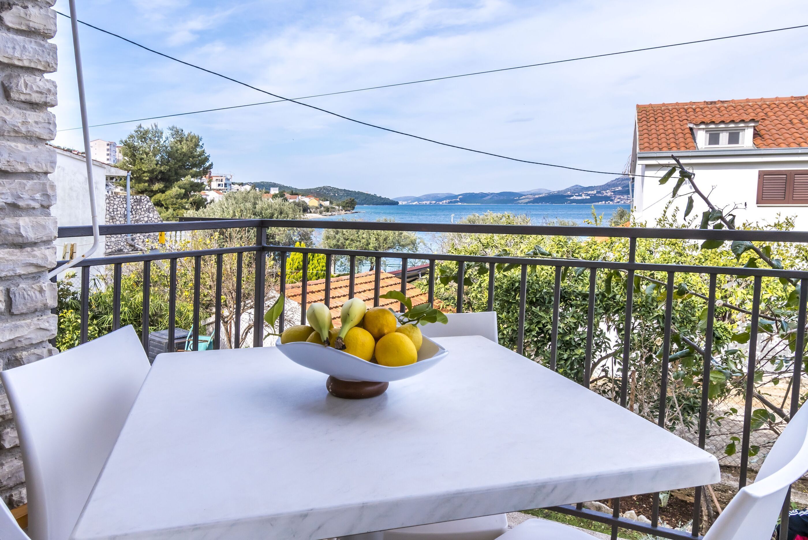 Un balcón con mesa y sillas, un cuenco de fruta y vistas al mar y colinas al distante.
