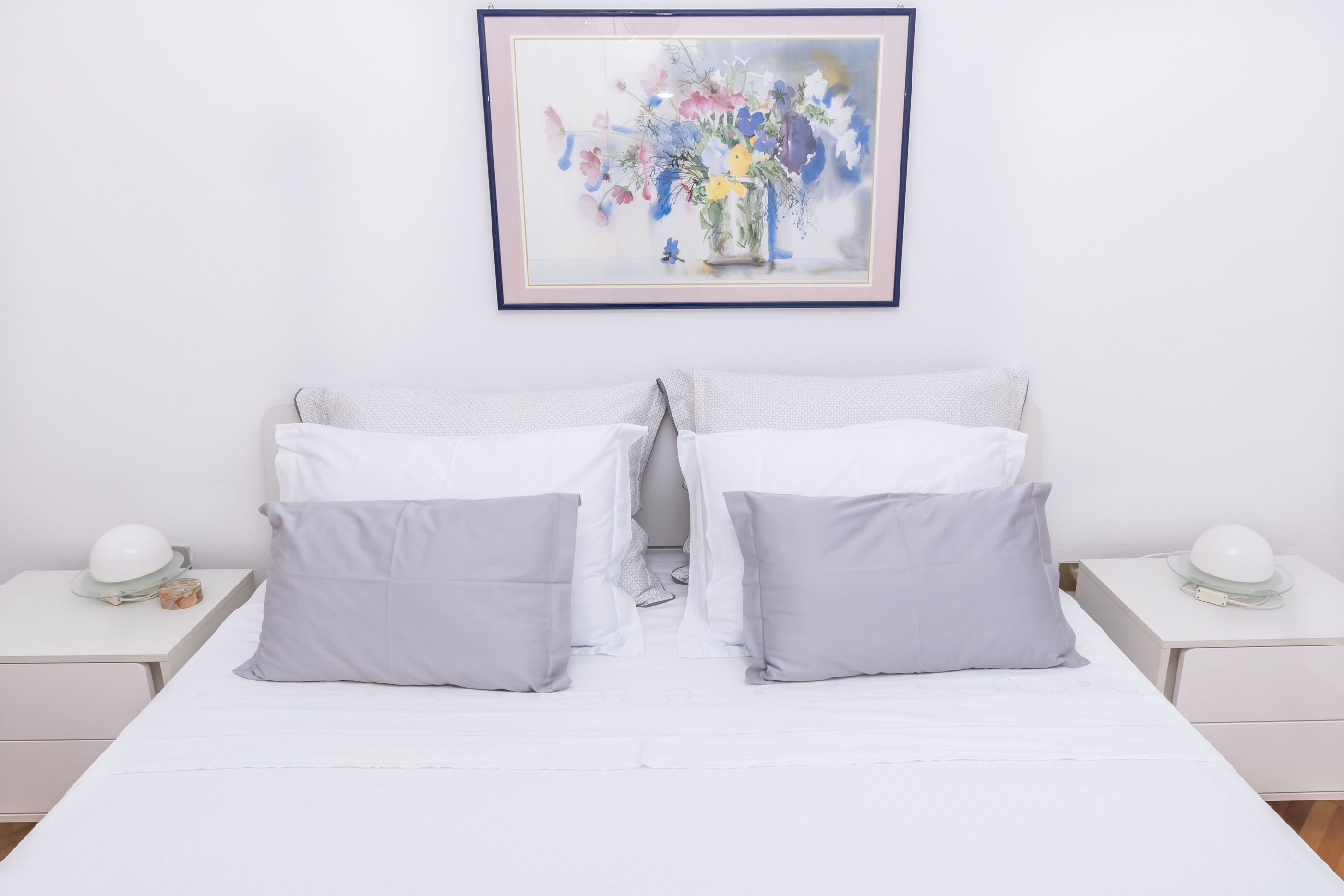 Un letto matrimoniale ordinato con biancheria bianca e due cuscini grigi, affiancato da due comodini bianchi con lampade e un quadro floreale incorniciato sopra.
