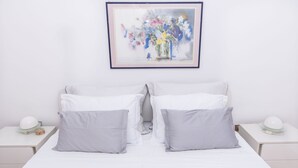 A neatly arranged double bed with white bedding and two gray pillows, flanked by white nightstands with lamps and a framed floral artwork above. - House Holiday House (Okrug)