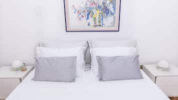 Un letto matrimoniale ordinato con biancheria bianca e due cuscini grigi, affiancato da due comodini bianchi con lampade e un quadro floreale incorniciato sopra.