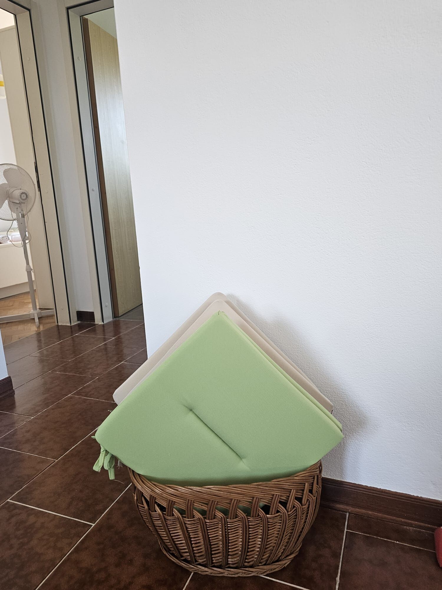 A wicker basket with a green cushion against a white wall, a fan in the background, and a tiled floor.
