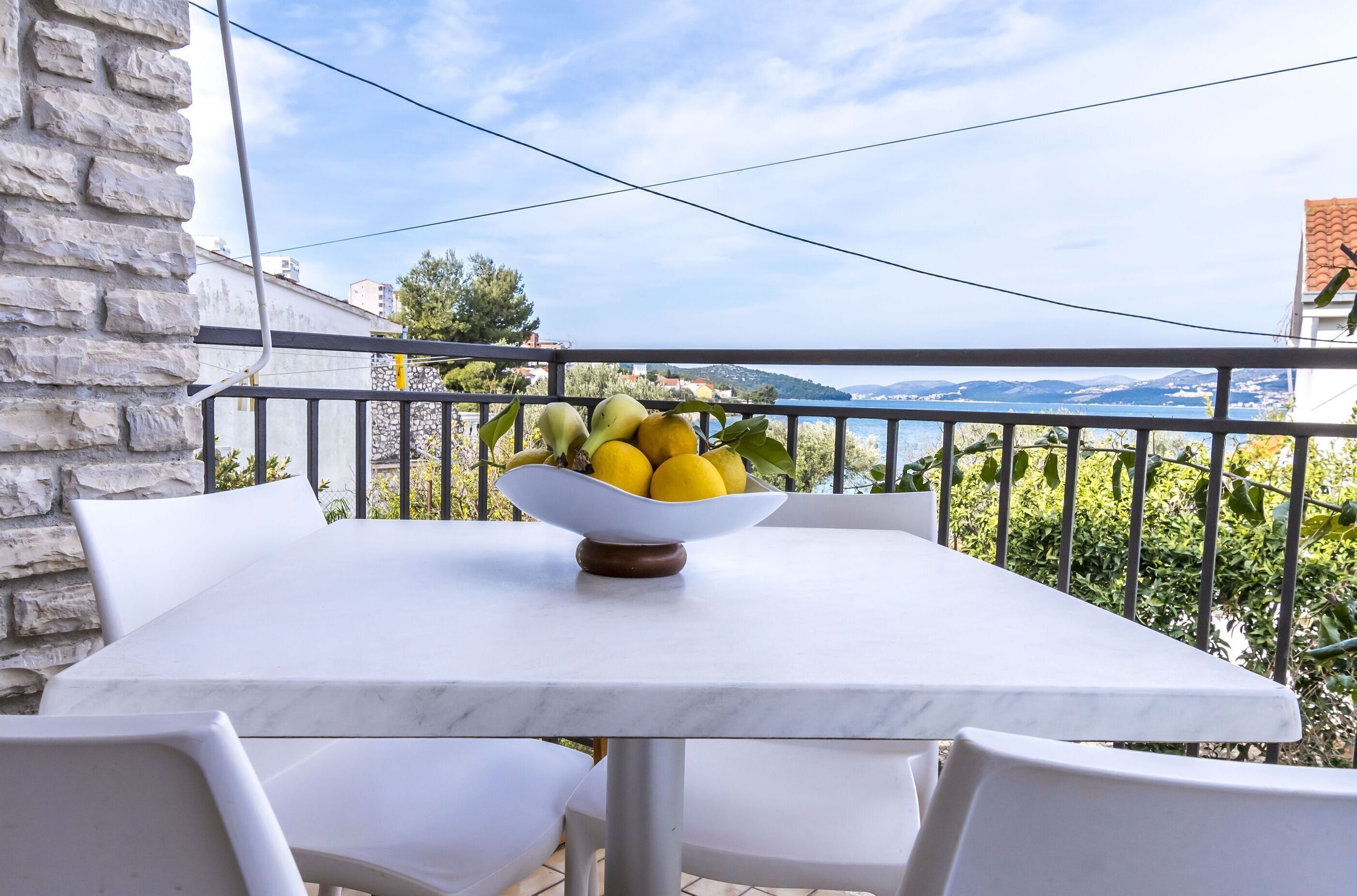 A balcony with a table and chairs, a bowl of fruit, and a view of the sea and hills.