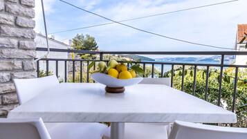 A balcony with a table and chairs, a bowl of fruit, and a view of the sea and hills.