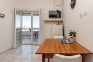 Een kamer met een houten tafel, witte stoelen, een aan de muur bevestigde televisie, een koelkast, een potplant en een schuifdeur met uitzicht op een buitenruimte.