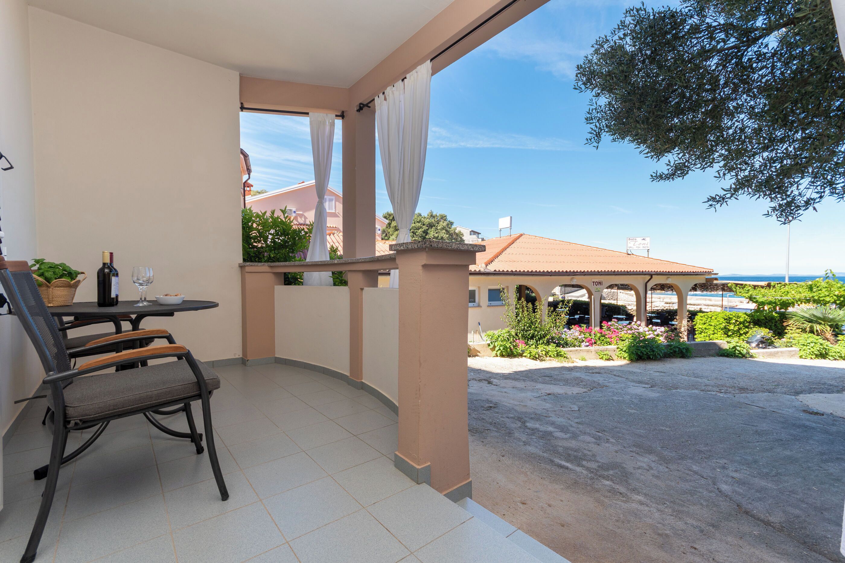 A balcony with a table and chairs, a bottle of wine, and a view of a building with a tiled roof.