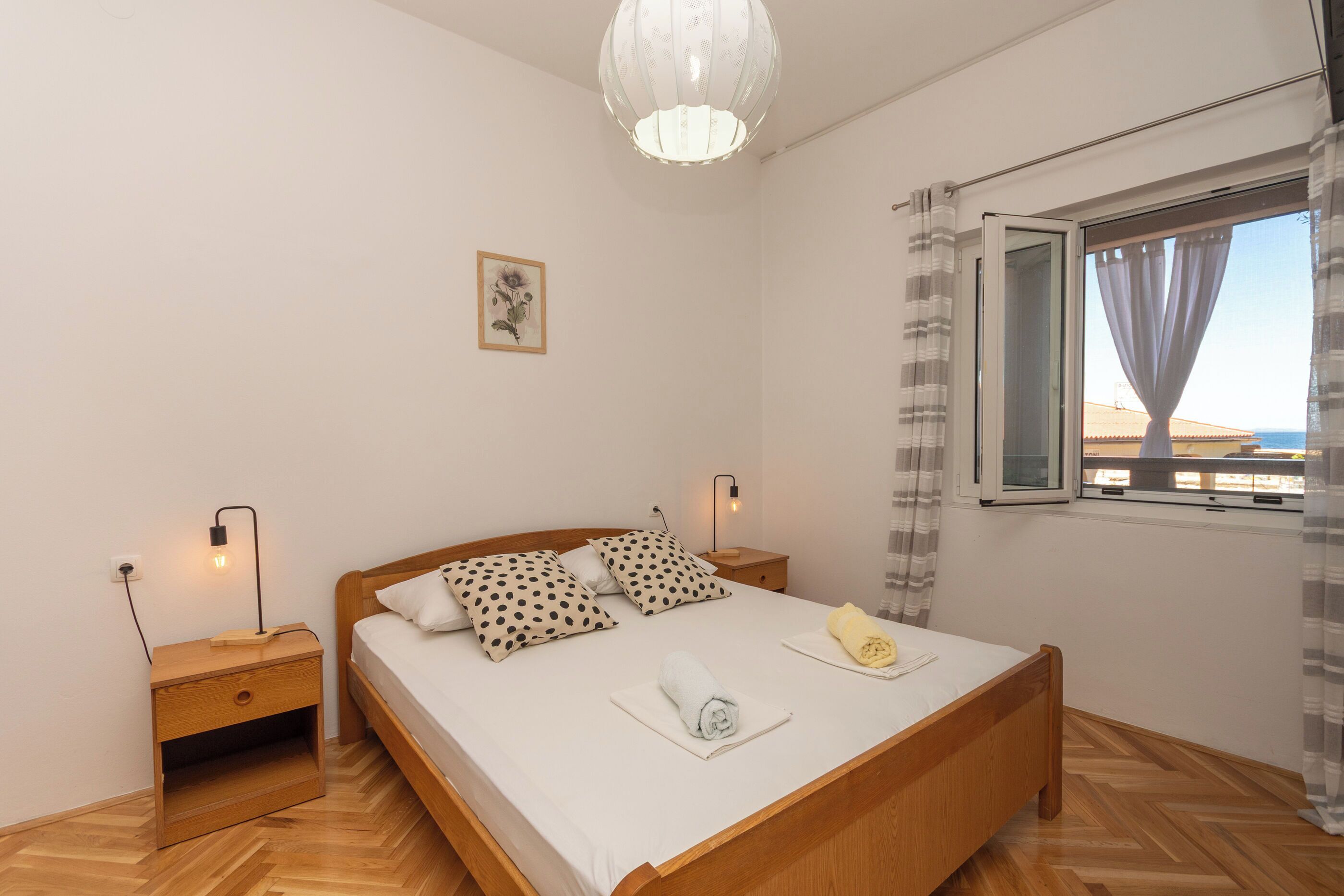 A bedroom with a wooden bed, a nightstand, a bedside lamp, a window with curtains, and a wall-mounted light fixture.