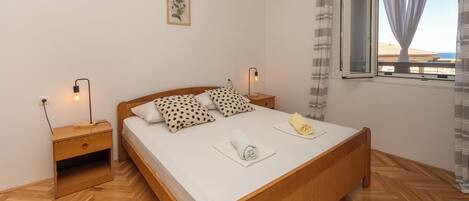 A bedroom with a wooden bed, a nightstand, a bedside lamp, a window with curtains, and a wall-mounted light fixture.