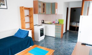 A compact room with a kitchenette, a bed with a blue cover, and a wooden table. - Apartments Seafront Oasis (Ston)