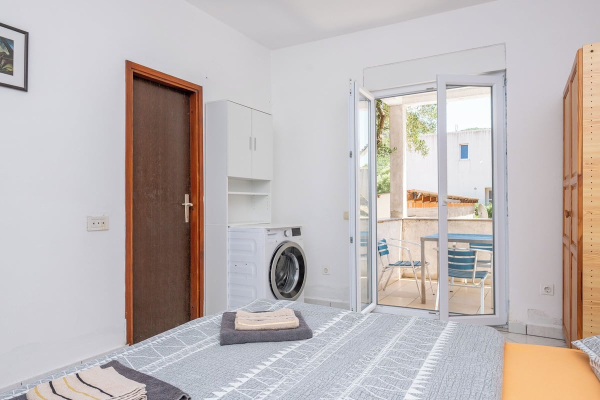 A bedroom with a bed, a washing machine, a wooden door, and a sliding glass door leading to an outdoor area with chairs and a table.